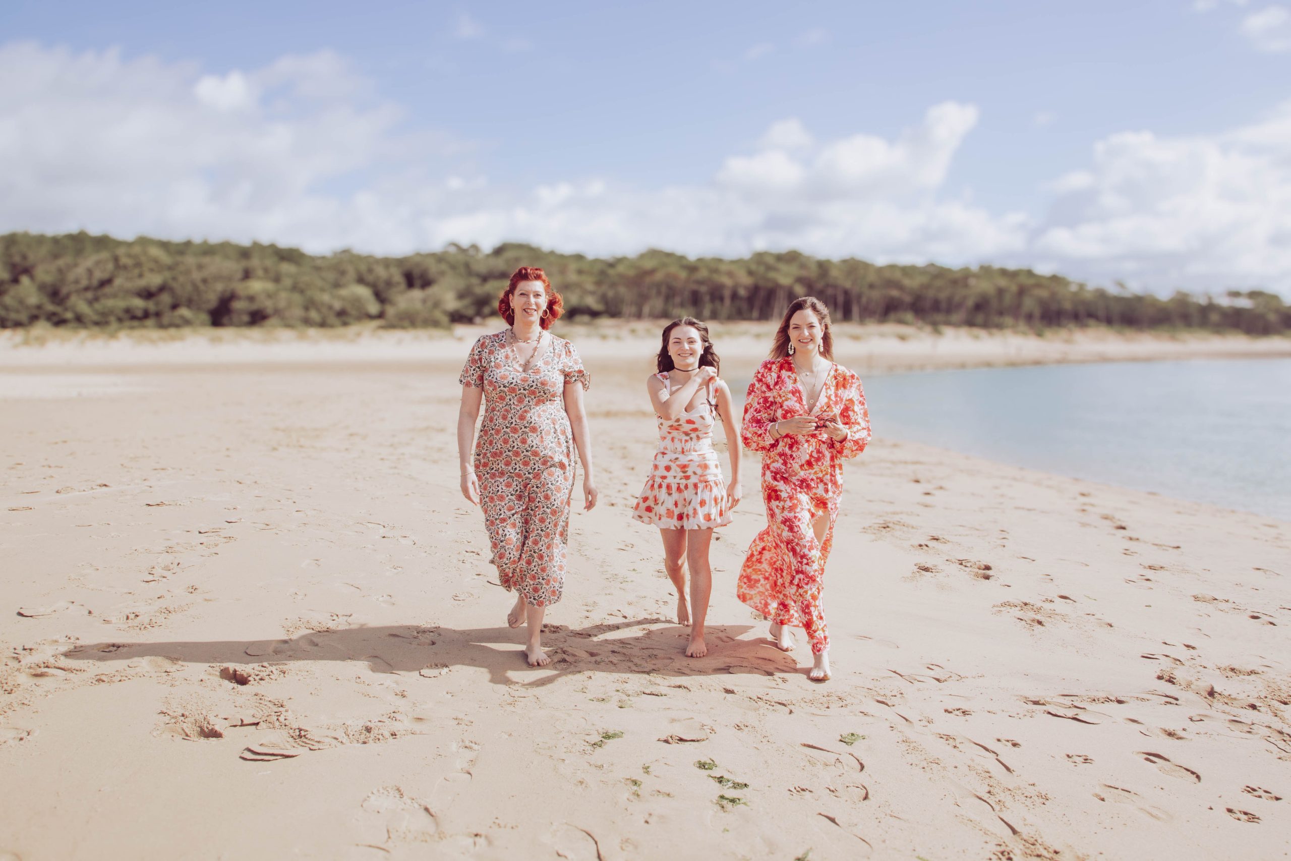 Colorimétrie Trois femmes marchant sur une plage ensoleillée, habillées de robes d'été à motifs floraux.
